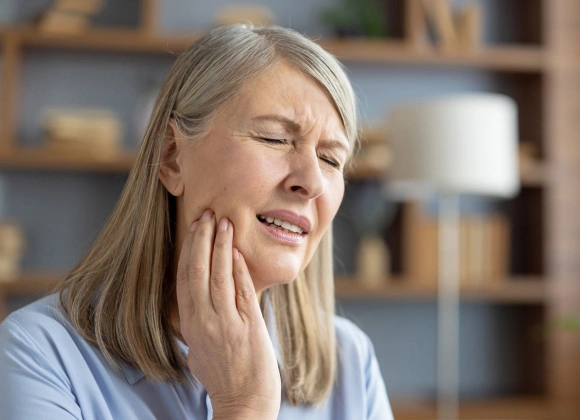 Woman experiencing tooth pain, holding jaw, in home setting with shelving background.