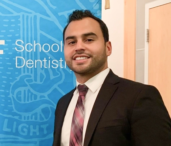 Professional male dentist in dark suit and pink tie smiling in modern dental office setting.