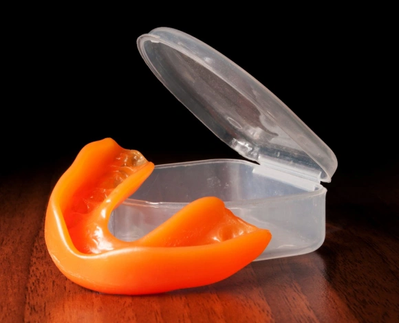 Clear orthodontic retainer case with orange mouthguard on wooden surface against black background.