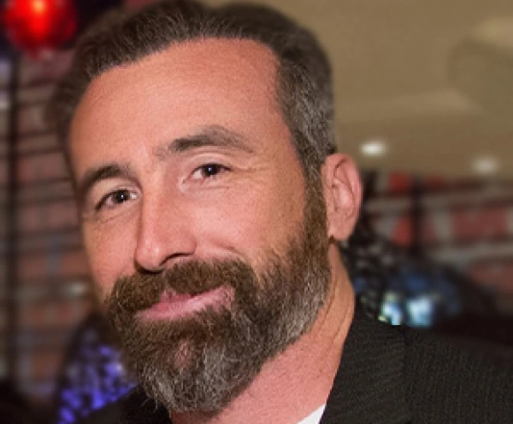 Professional headshot of bearded man in dental office setting with red light visible in background.