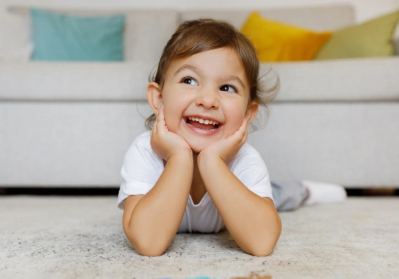 Happy toddler smiling on carpet with colorful pillows on couch behind.
