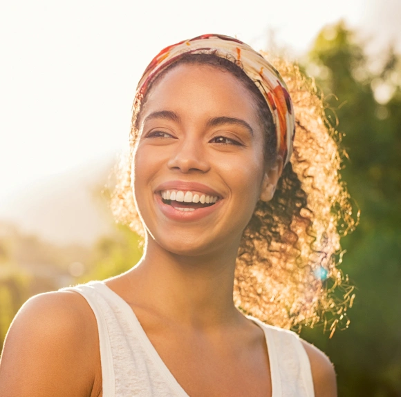 Woman smiling outdoors at sunset wearing white top and colorful headwrap.