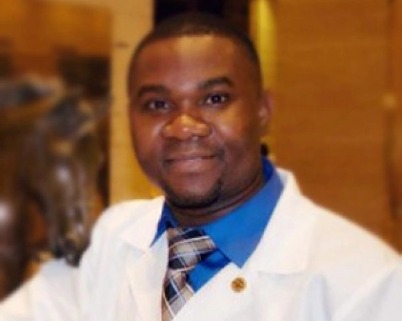 Professional headshot of male dentist in white coat and blue tie smiling at camera indoors.
