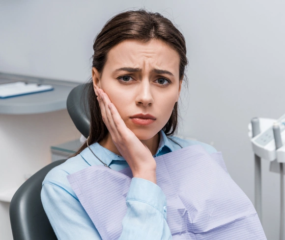 Woman in dental chair holding cheek, expressing discomfort or tooth pain in dental office.