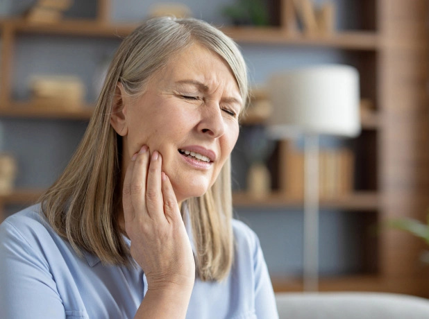 Middle-aged woman touching her cheek in discomfort, dental office setting with shelves in background.