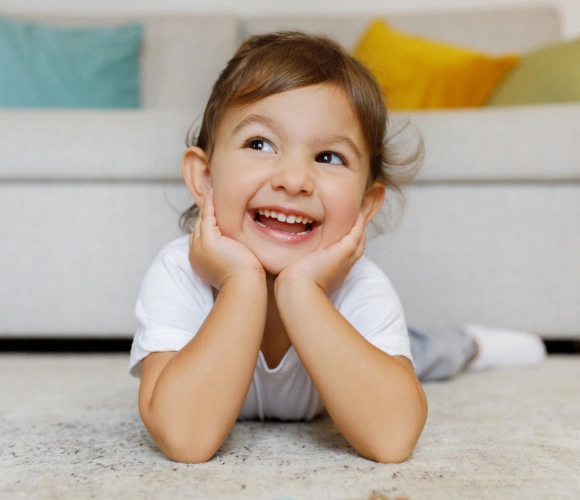 Happy toddler lying on carpet, smiling with hands on face in bright modern living room.
