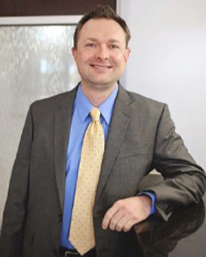 Professional man in gray blazer and blue shirt smiling in office setting.