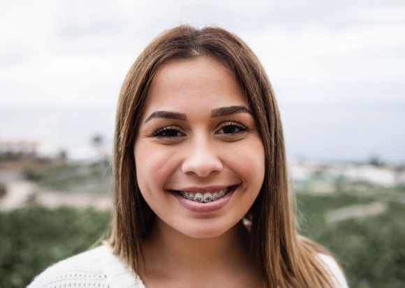 Young woman smiling at camera outdoors with landscape background, dental practice portrait.