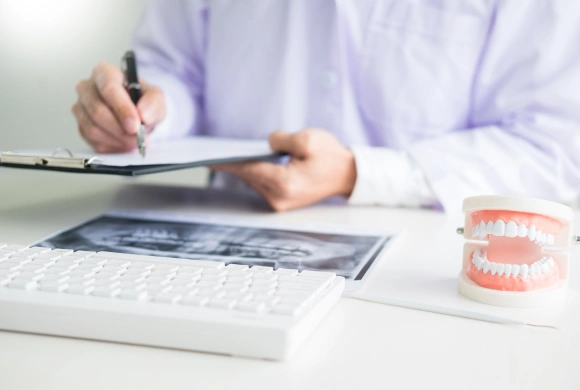 Dentist writing notes at desk with dental x-ray and teeth model displayed.