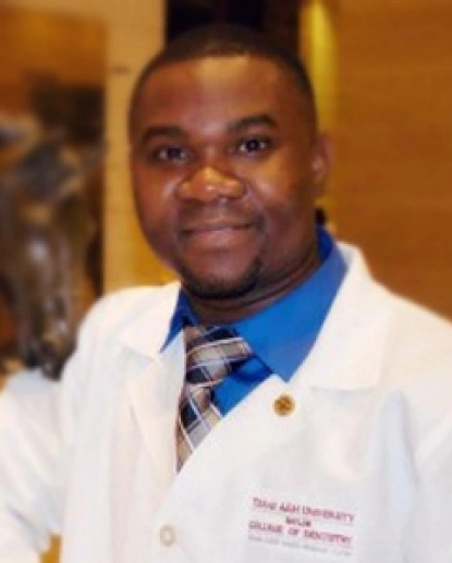 Male dentist in white coat and blue tie smiling at camera in clinical setting.