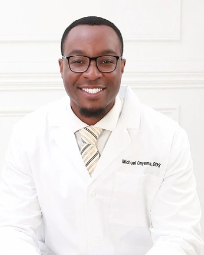 Male dentist wearing white coat and glasses smiling at camera in professional dental office setting.