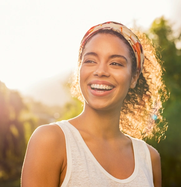 Happy woman smiling outdoors in sunlight, wearing white tank top and headwrap.
