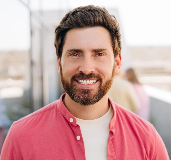 Professional headshot of smiling man in pink shirt against blurred urban rooftop background.