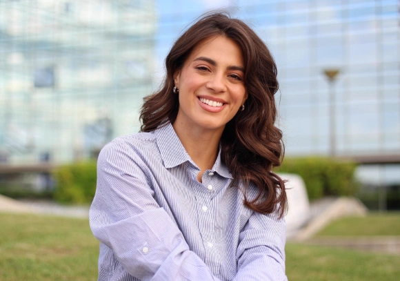 Smiling woman in lavender shirt outdoors with modern buildings and green lawn in background.