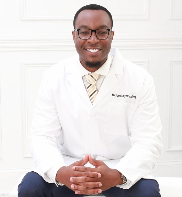 Professional headshot of male dentist in white coat and glasses against white clinical background.