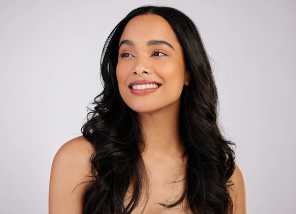 Woman with long black wavy hair smiling against white background, dental practice portrait.
