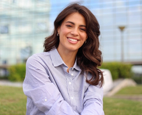 Professional woman smiling outdoors in business casual attire, modern office buildings visible in background.