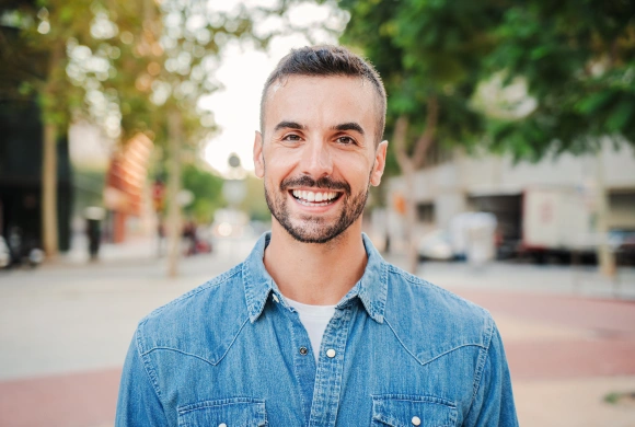 Smiling man in denim shirt standing outdoors on city street during daytime.