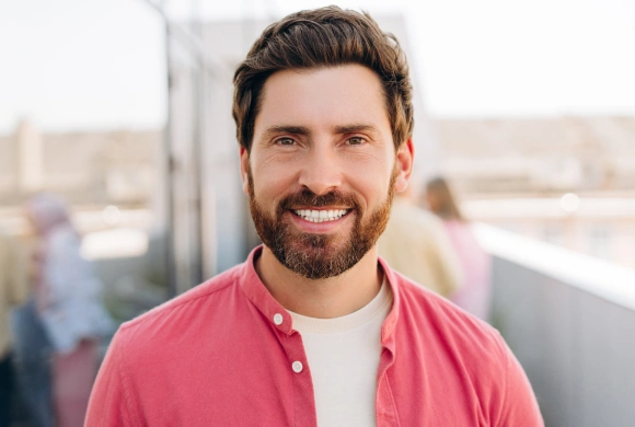 Smiling man in pink shirt standing on rooftop with urban background.