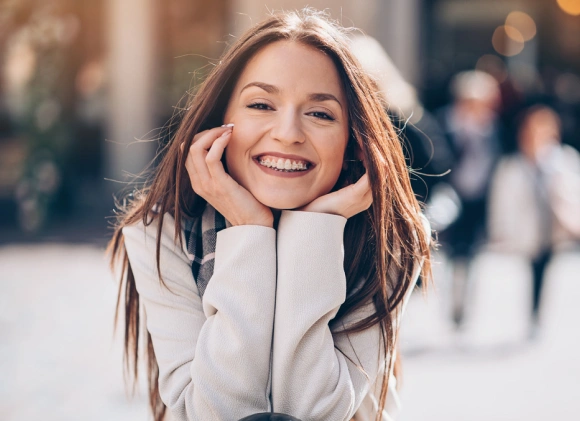 Young woman smiling outdoors, wearing light sweater, hands near face in casual pose.